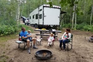 Family sitting by fire behind RV campsite.