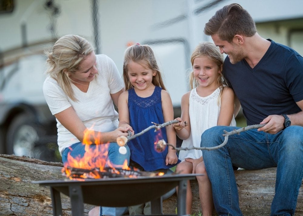 Family roasting marshmallows around campfire in front of RV.