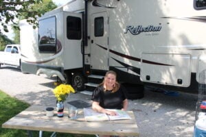 Woman sitting in shade beside RV on sunny day. 