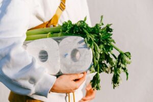 Close up of woman's arm holding groceries, including toilet paper and vegetables. 