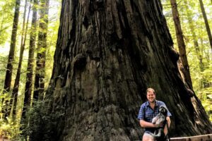 Man holding dog sitting in front of Redwood tree.