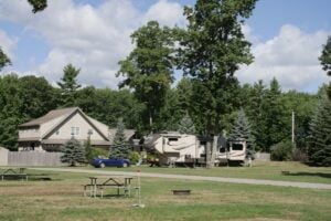 RV parked at campsite with tall trees and office building in background.