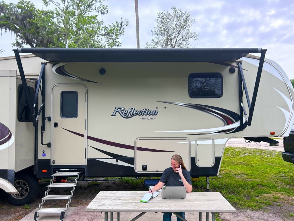 Woman at RV campsite picnic table with phone, computer and notepad.