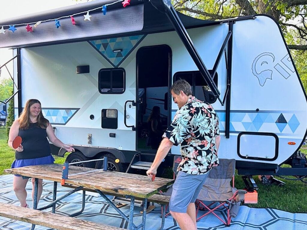 Man and woman playing table tennis on campsite picnic table.